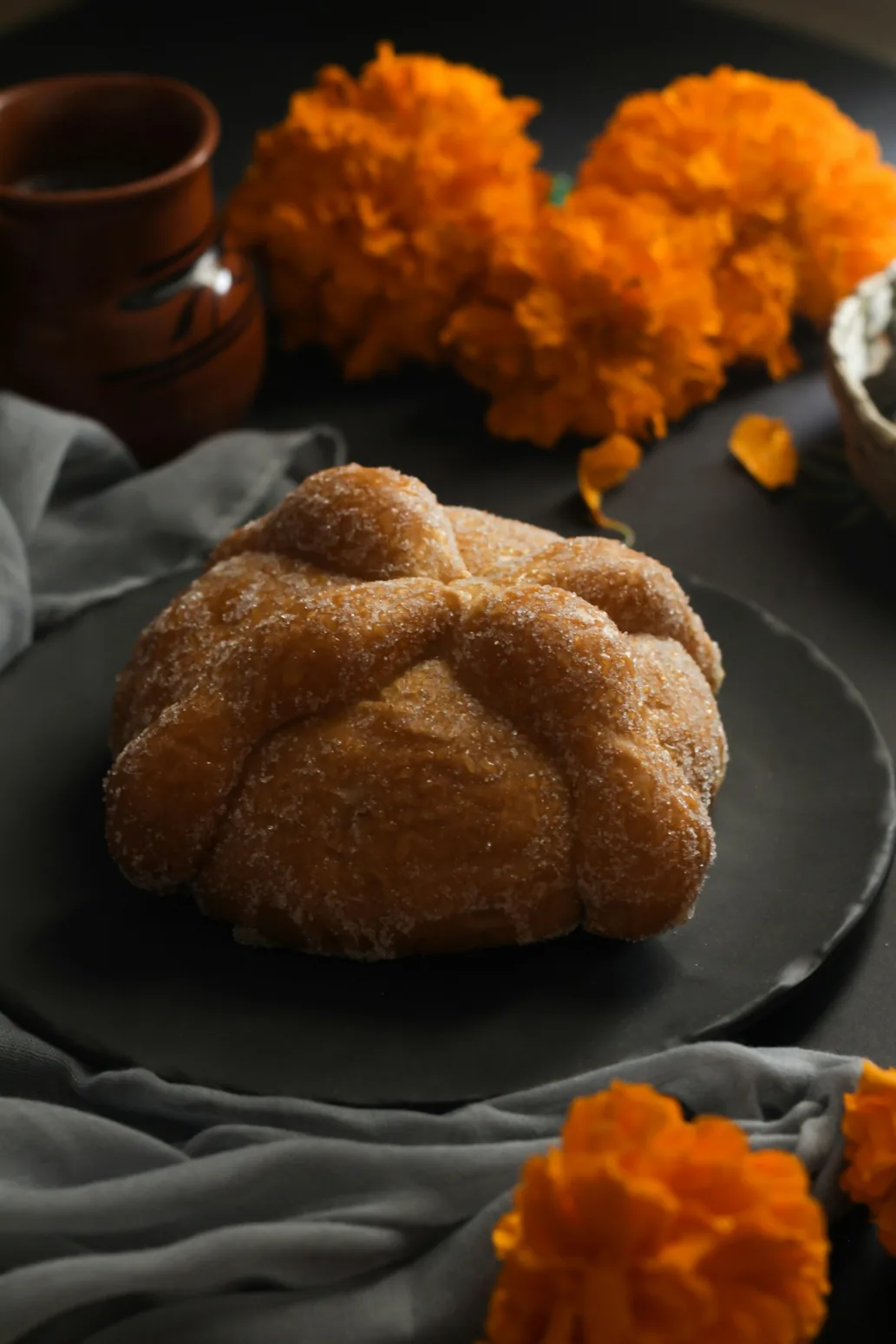 Tradiční sladký pan de muerto na tmavém talíři s oranžovými květy cempasúchil