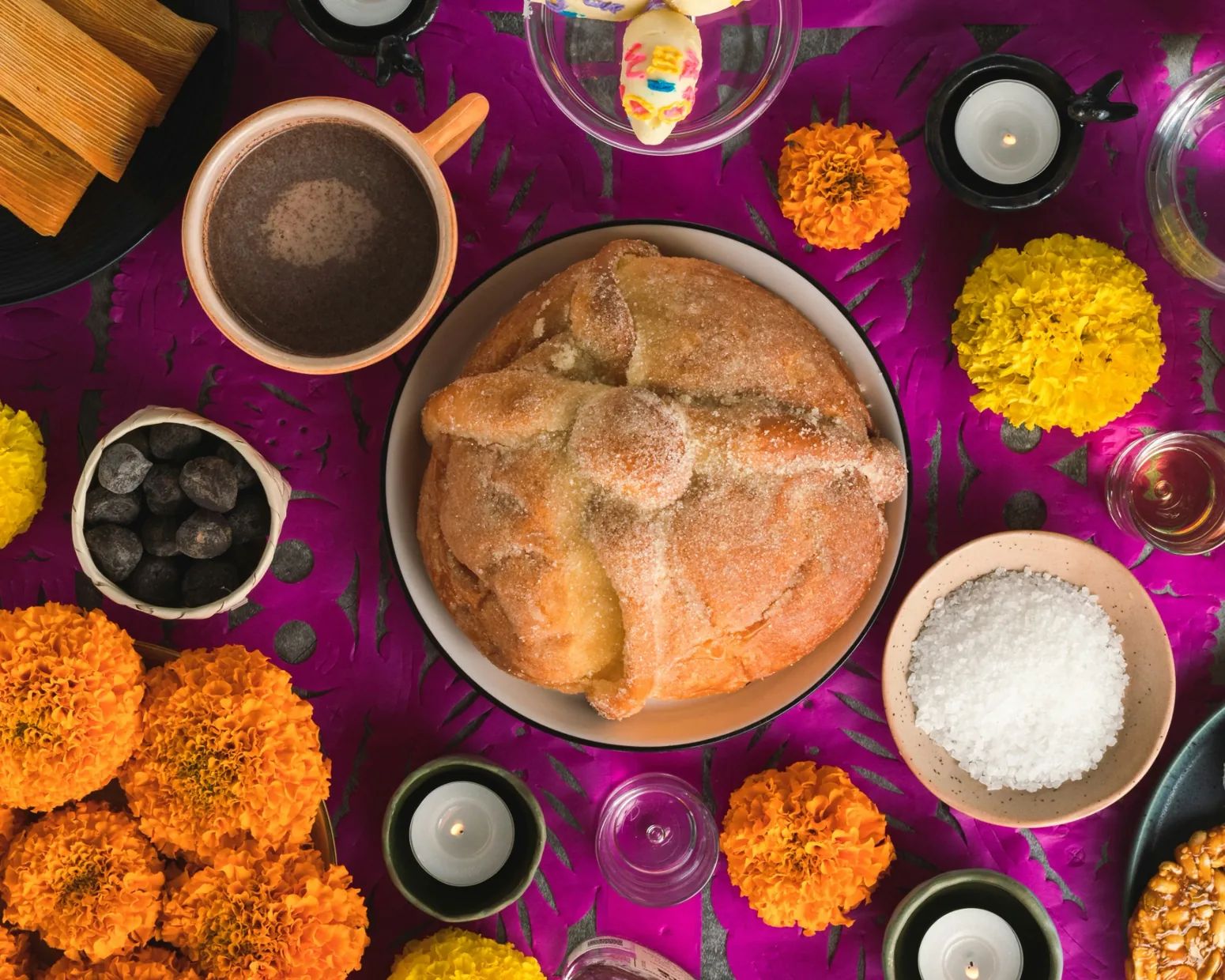 Pan de muerto na barevném oltářním ubruse mezi kakaem, svíčkami a květy cempasúchil