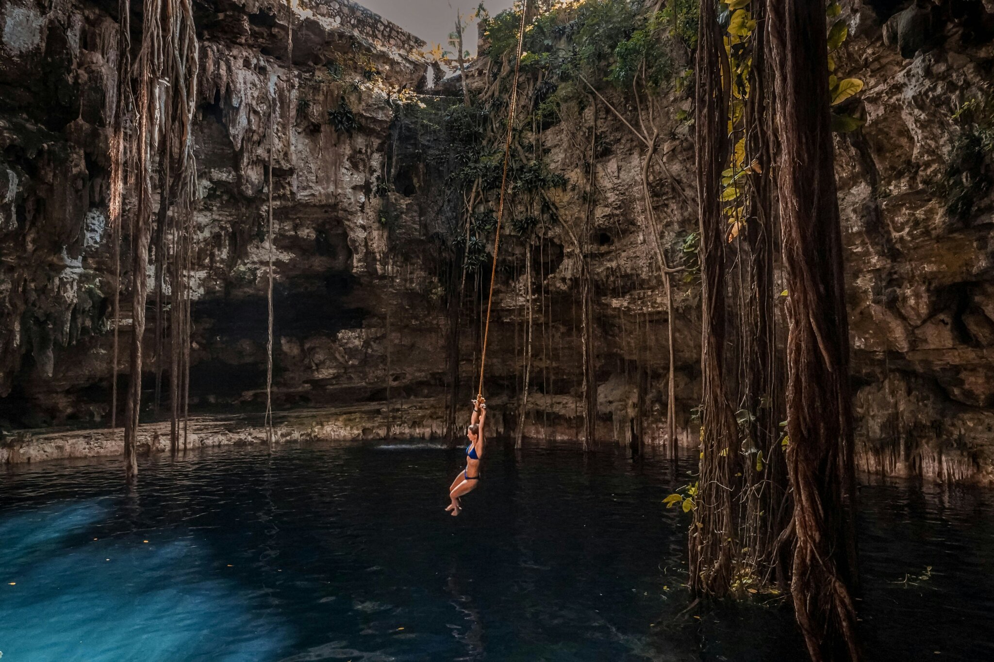 Otevřená cenota na Yucatánu s visícími kořeny, temně modrou vodou a turistkou skákající do cenoty na laně