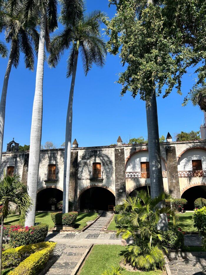 Pohled na zahradu historického místa Hacienda Vista Hermosa v Mexiku se zelenými palmami a architektonickými detaily budovy v pozadí, jasná modrá obloha.