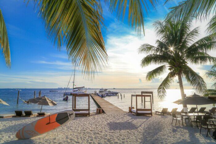 Pláž na ostrově Isla Mujeres u poloostrova Yucatan v Mexiku s bílým pískem, palmami, lehátky a molem vedoucím k jachtám při západu slunce